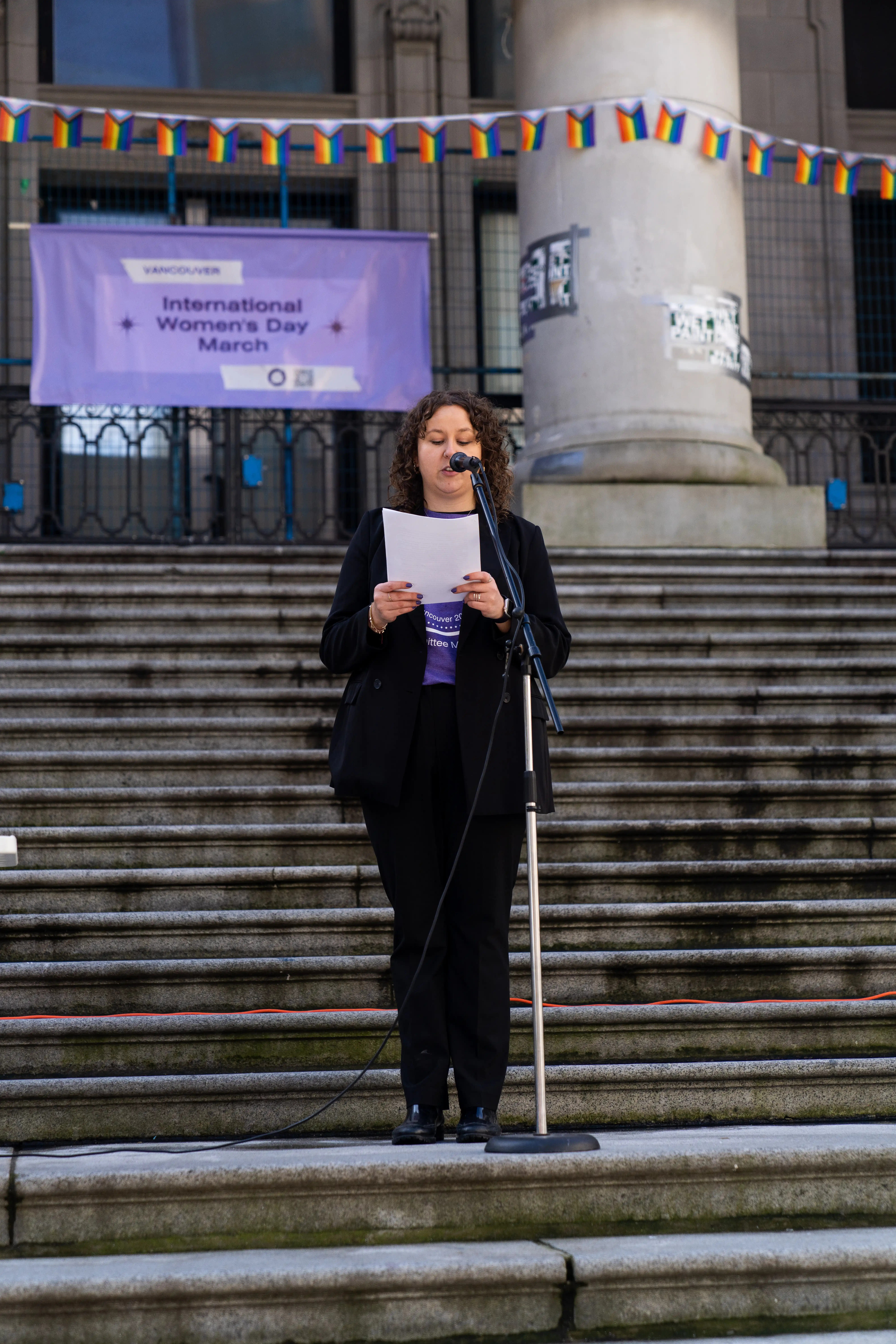 This photo is of the committee chair, at the 2023 Vancouver International Women’s Day march. She is standing on a large platform of stone stairs. She is standing in front of a microphone, reading off of paper to an audience. Behind her, is a large purple banner that reads International Women’s Day. She is wearing a black pant suit, with a purple shirt underneath. 

                    