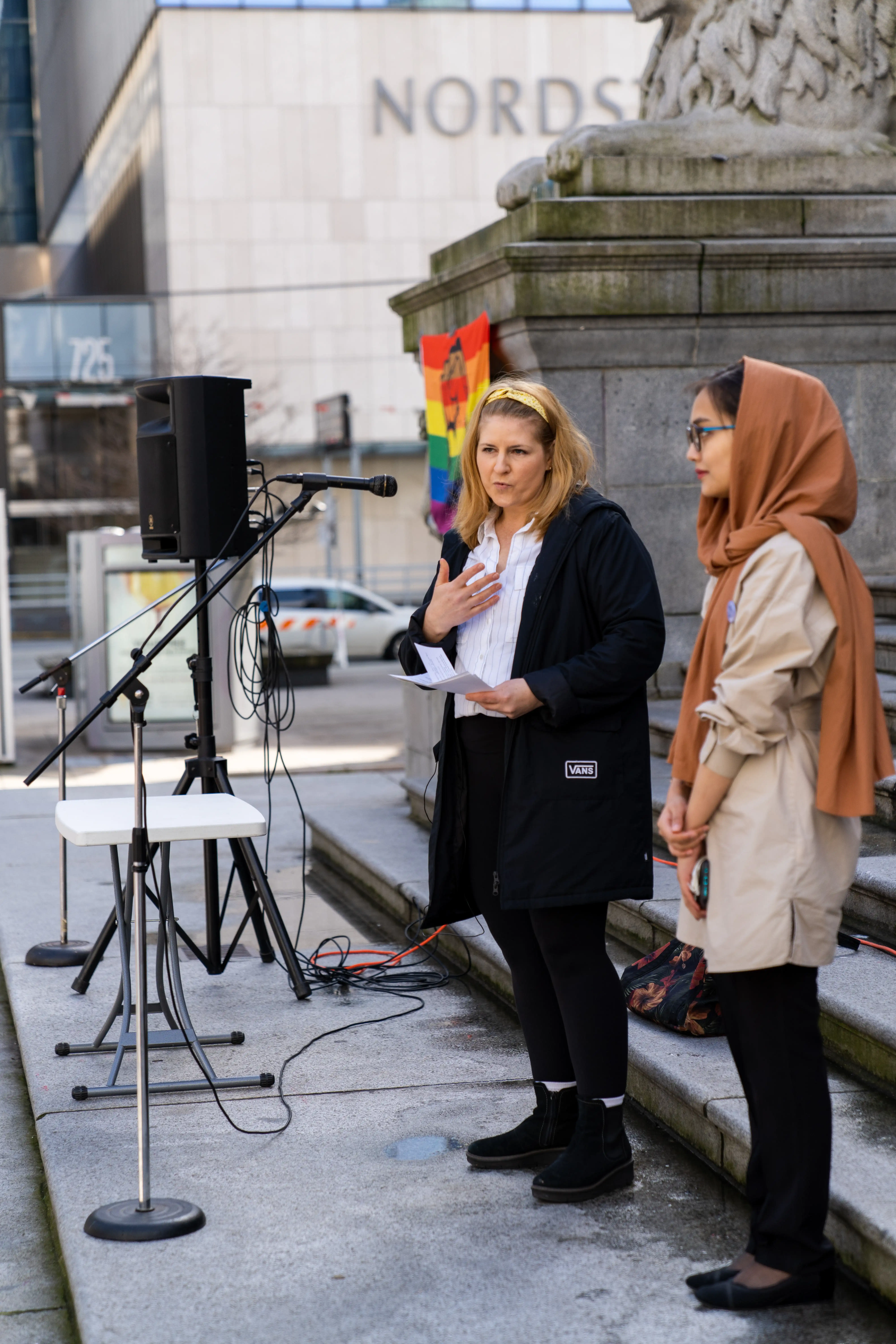 This is a photo of two of the speakers at the 2023 Vancouver International Women’s Day march. The two speakers are standing together on a large stone platform. They are both holding papers, reading off of them into the microphone. 