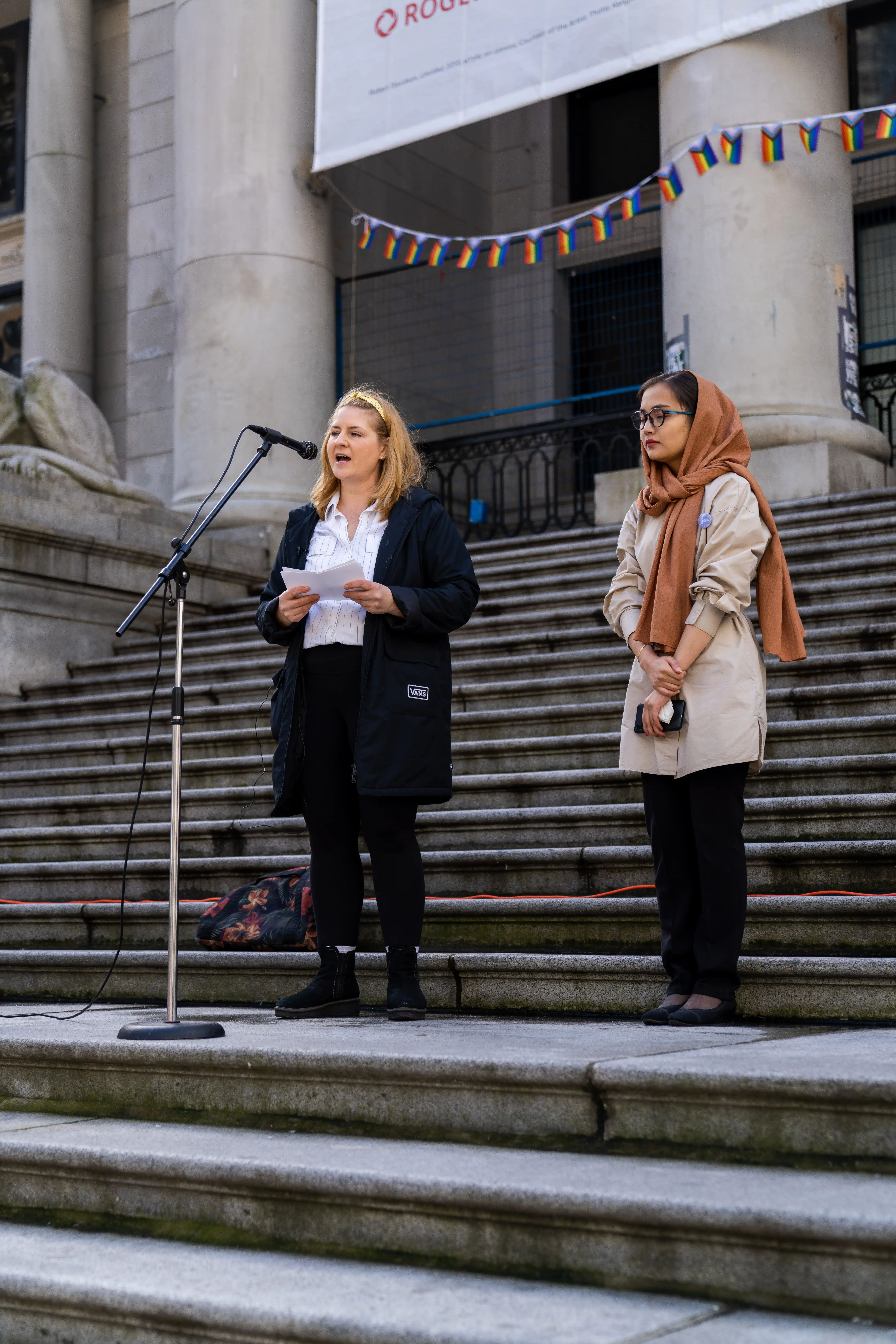 This is an image of Lauryn Oates and Sahar Maqsoodi, speaking to the crowd. They are speaking into a microphone on large stone stairs outside. 