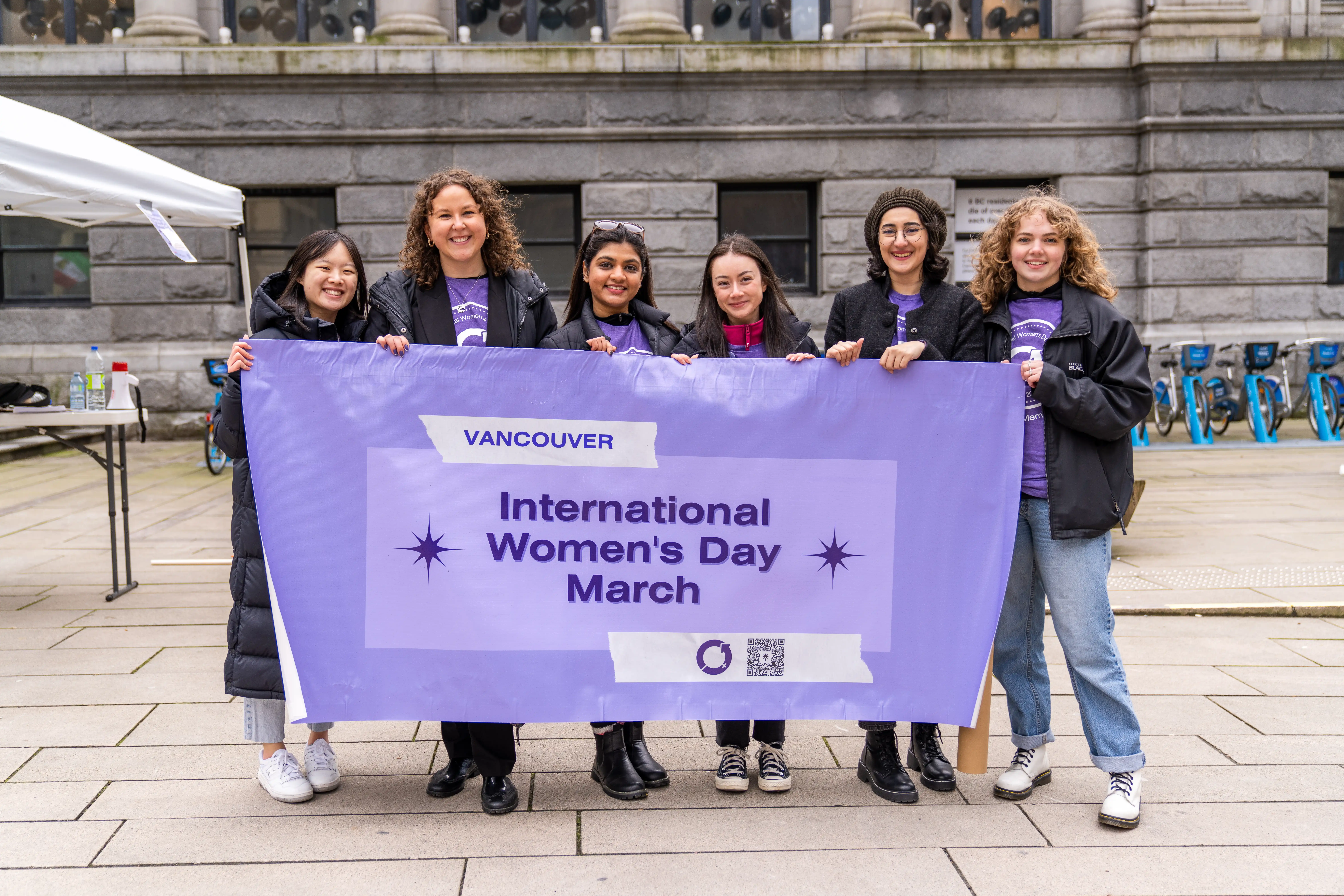 This is a photo of the 2023 event organizers. The six people are standing behind a large banner they are holding. The banner reads “international women’s day march.” 