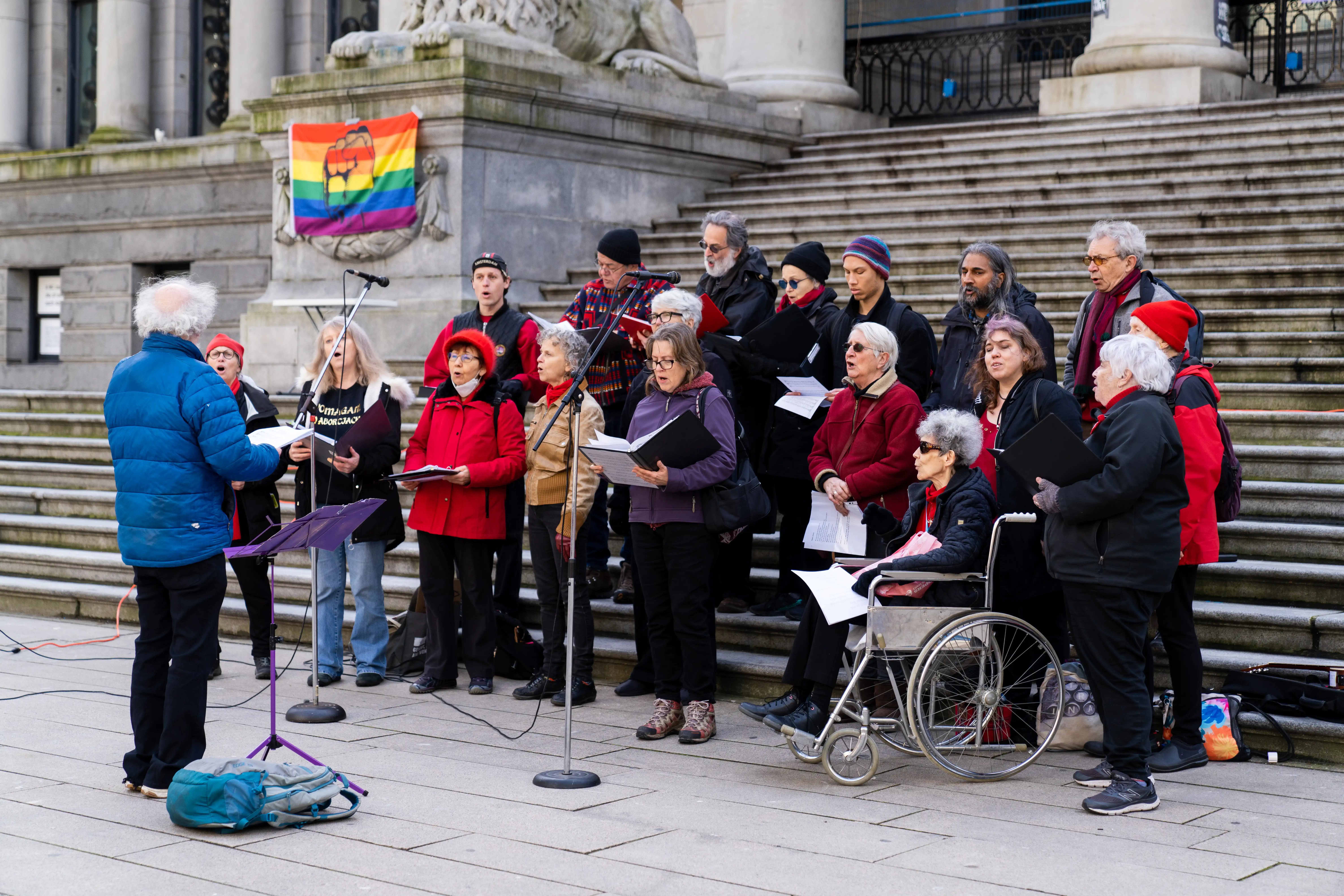 This is a photo of the Solidarity Notes, singing at the 2023 event. They are standing in two rows on large stone stairs. There are multiple microphones in front of them