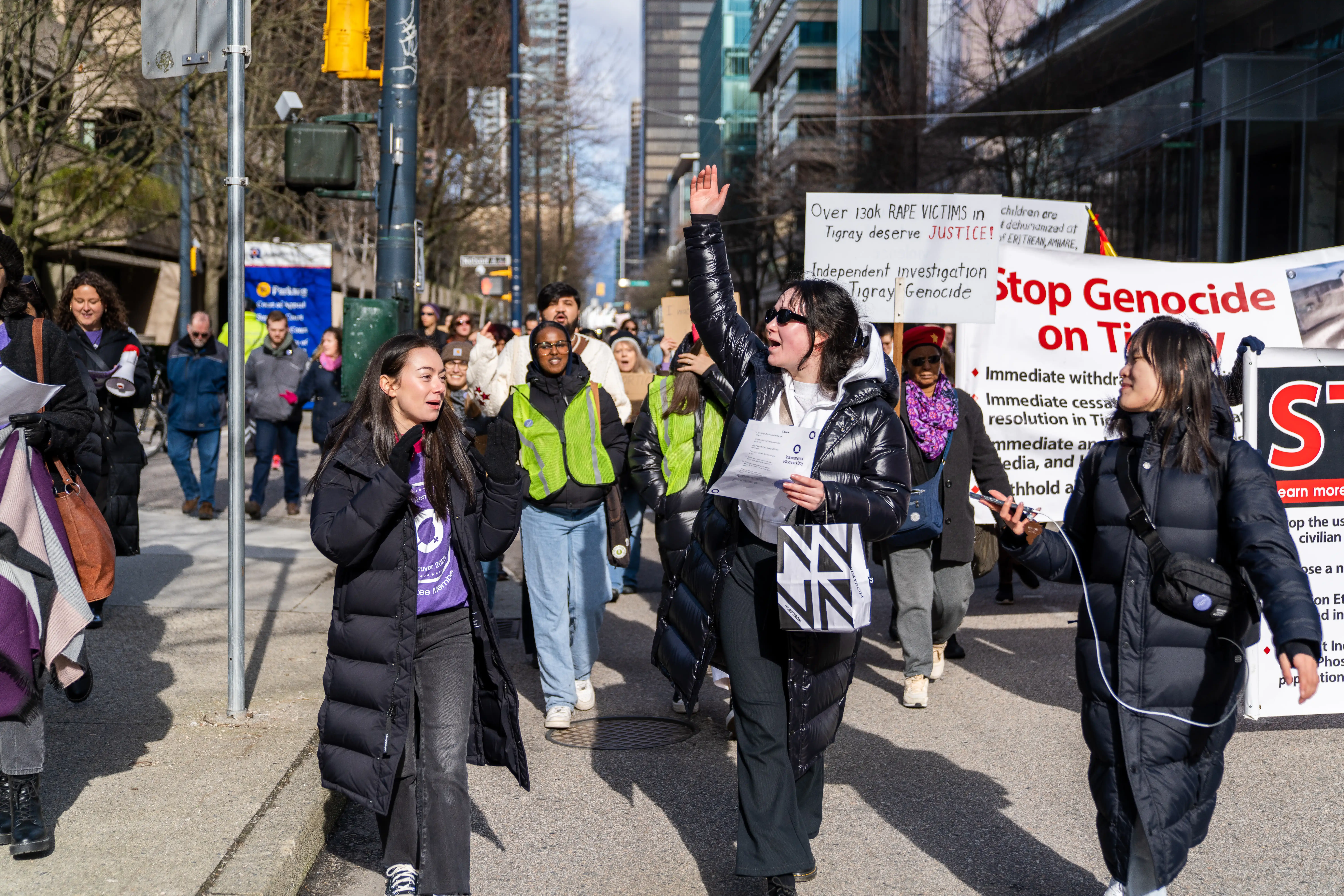 This is a photo of the march participants from the 2023 Vancouver event. The marchers are walking towards the camera, dressed in warm winter coats. Many are holding signs, clapping, and smiling.
