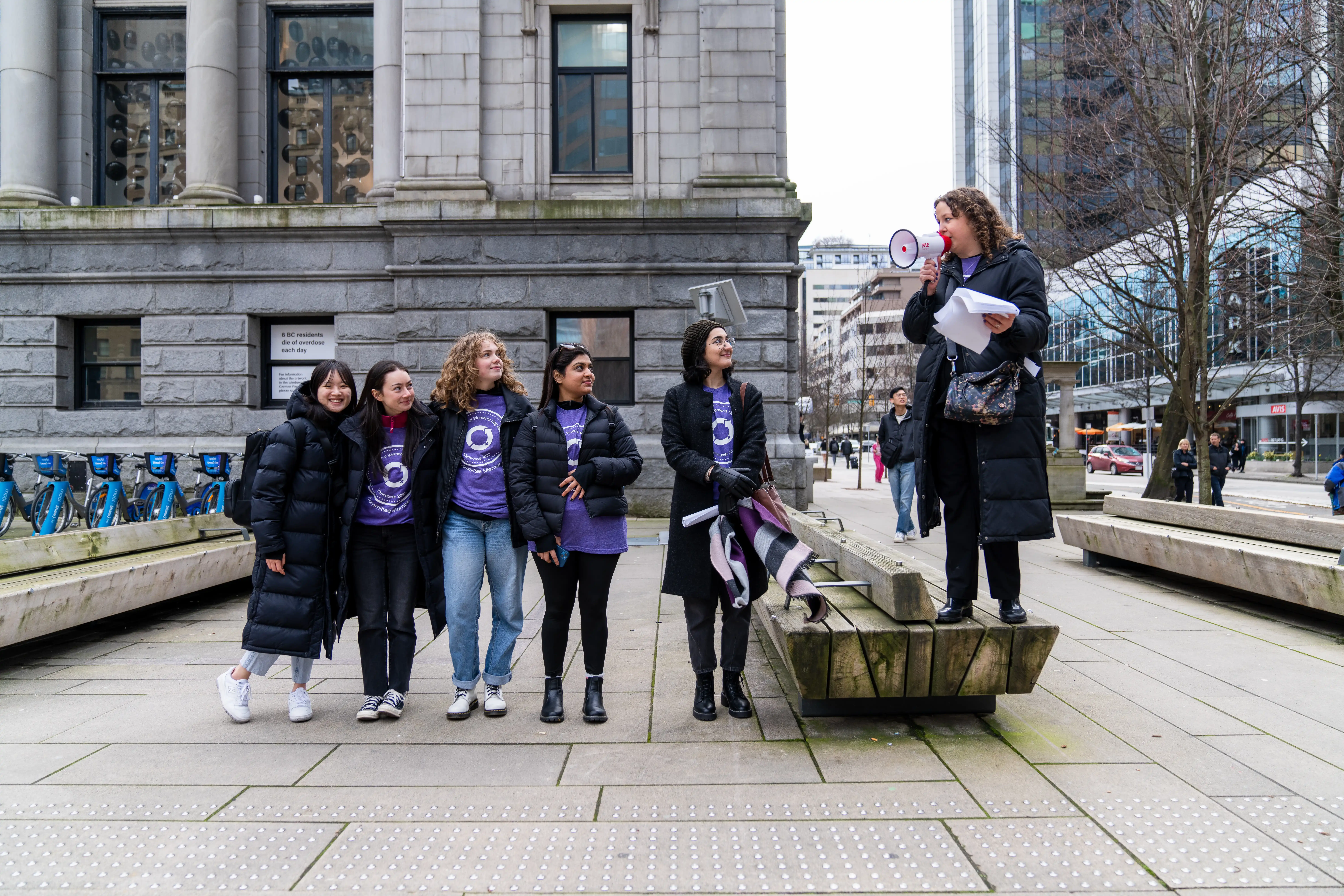 This is a photo of the 2023 organizing committee. The committee chair, Yasmin, is standing on a bench with a megaphone, speaking to the crowd. The rest of the organizing committee is standing next to her. They are arm-in-arm. 